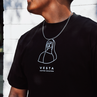 Person wearing a black t-shirt with Vesta Coffee Roasters logo against a white brick wall.