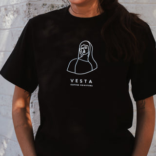 Person wearing a black t-shirt with 'Vesta Coffee Roasters' logo against a gray brick wall.